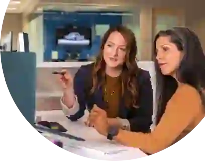 The picture shows two women working at their computers in the office and discussing something together. Their gaze is directed to the left.