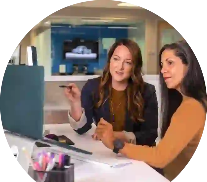 The picture shows two women working at their computers in the office and discussing something together. Their gaze is directed to the left.
